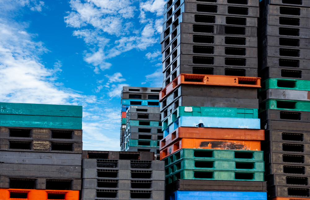 Tall stacks of plastic pallets in grey, green, blue and orange under a blue sky.